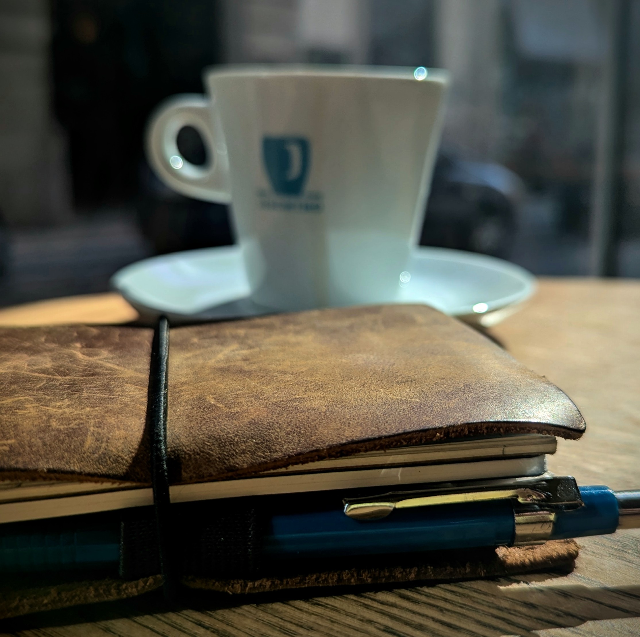 Uma xícara de café e um caderno de notas com aspecto antigo sobre uma mesa de um café. A mesa está próxima da janela e está sendo iluminada pelos primeiros raios de sol.
