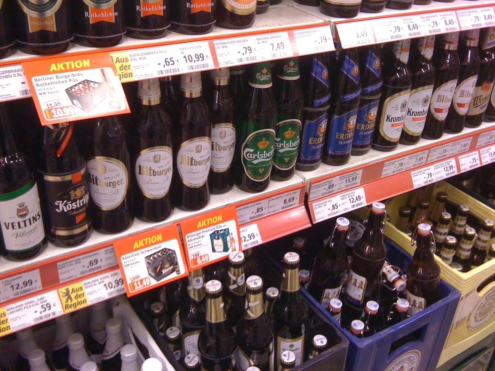 Bottles of various brands of beer are displayed on grocery store shelves, along with pricing and promotional labels.