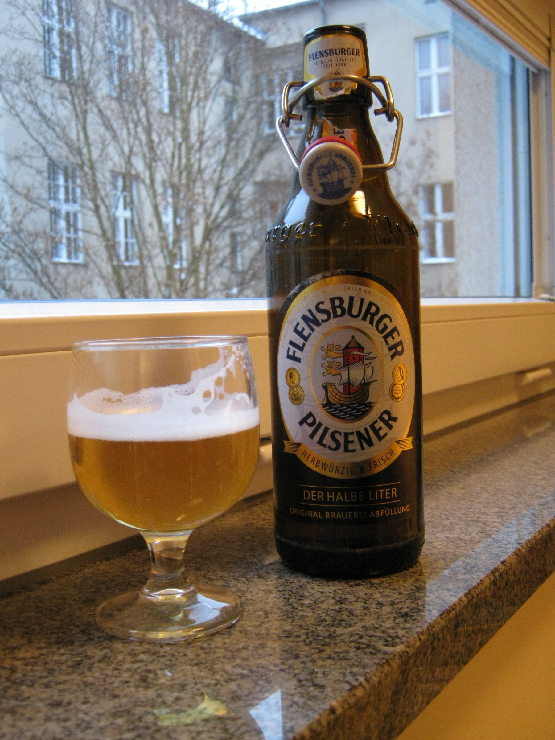 A Flensburger Pilsener bottle is placed next to a half-filled glass of beer on a windowsill.