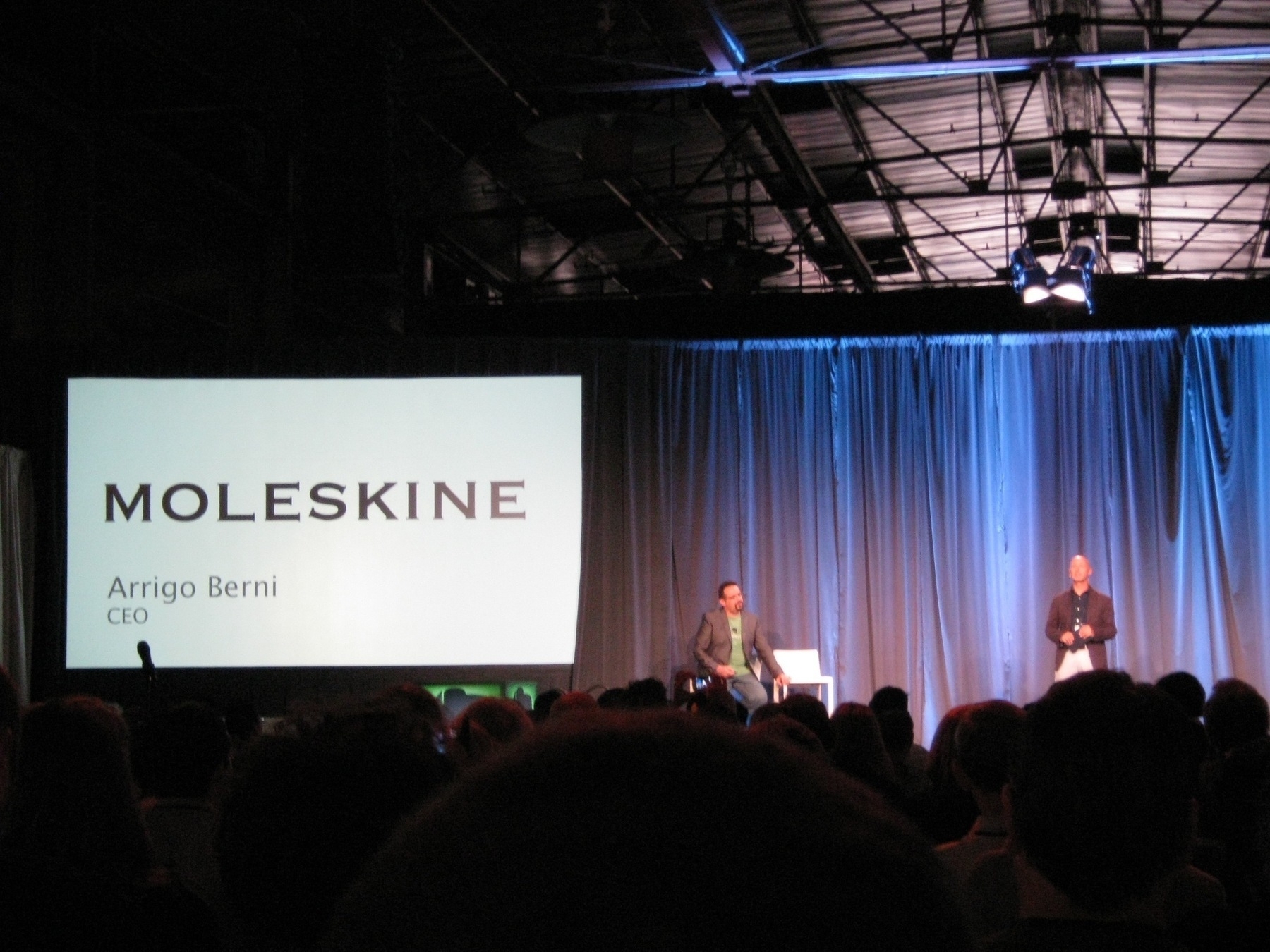 A speaker stands on stage with a large screen displaying the Moleskine logo and the name Arrigo Berni, CEO, in front of an audience.