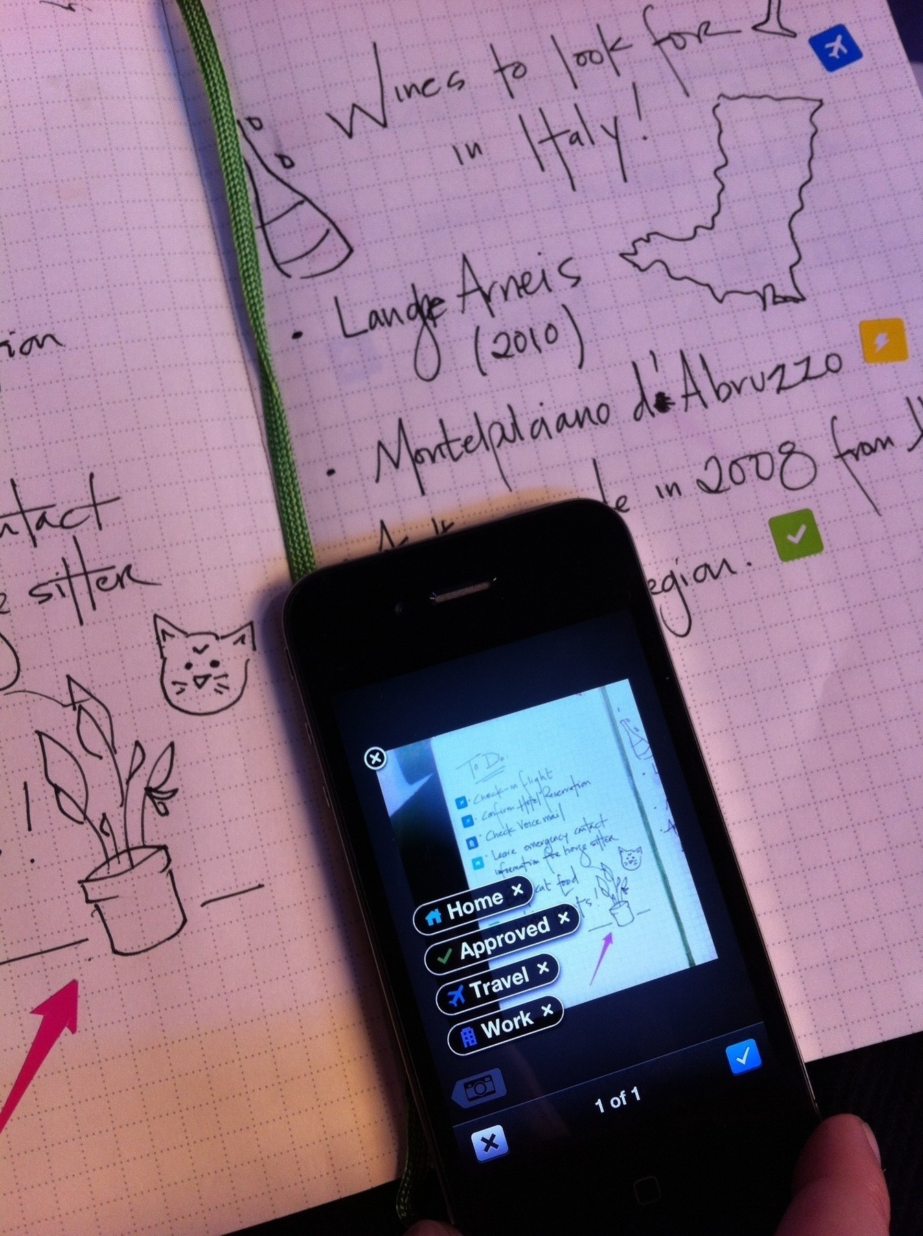 A smartphone captures notes from a notebook featuring handwritten lists and sketches, including a cat and a potted plant.