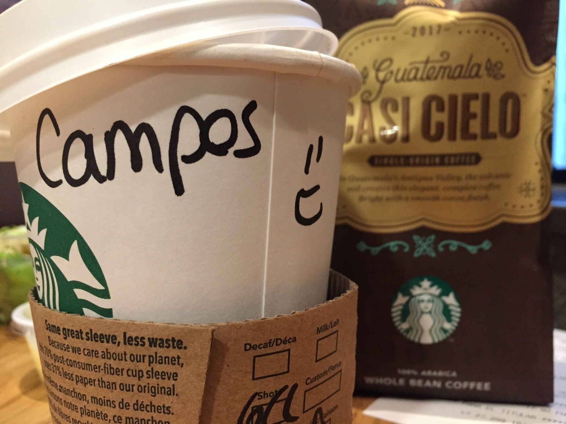 A Starbucks cup with Campos and a smiley face written on it is next to a bag of Guatemala Casi Cielo whole bean coffee.