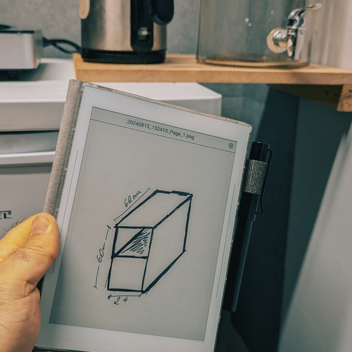 A hand is holding a Supernote device displaying a technical sketch of a rectangular object with dimensions, while a kettle and other kitchenette items are visible in the background.