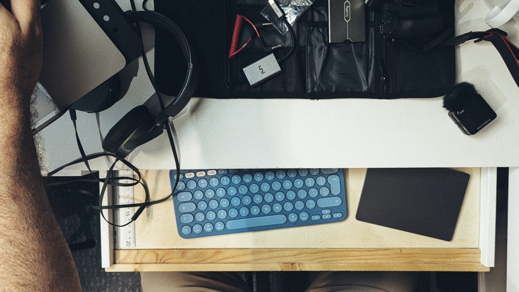 A person is sitting at a cluttered desk with various electronic devices, including a keyboard, headphones, and other tech accessories in view.