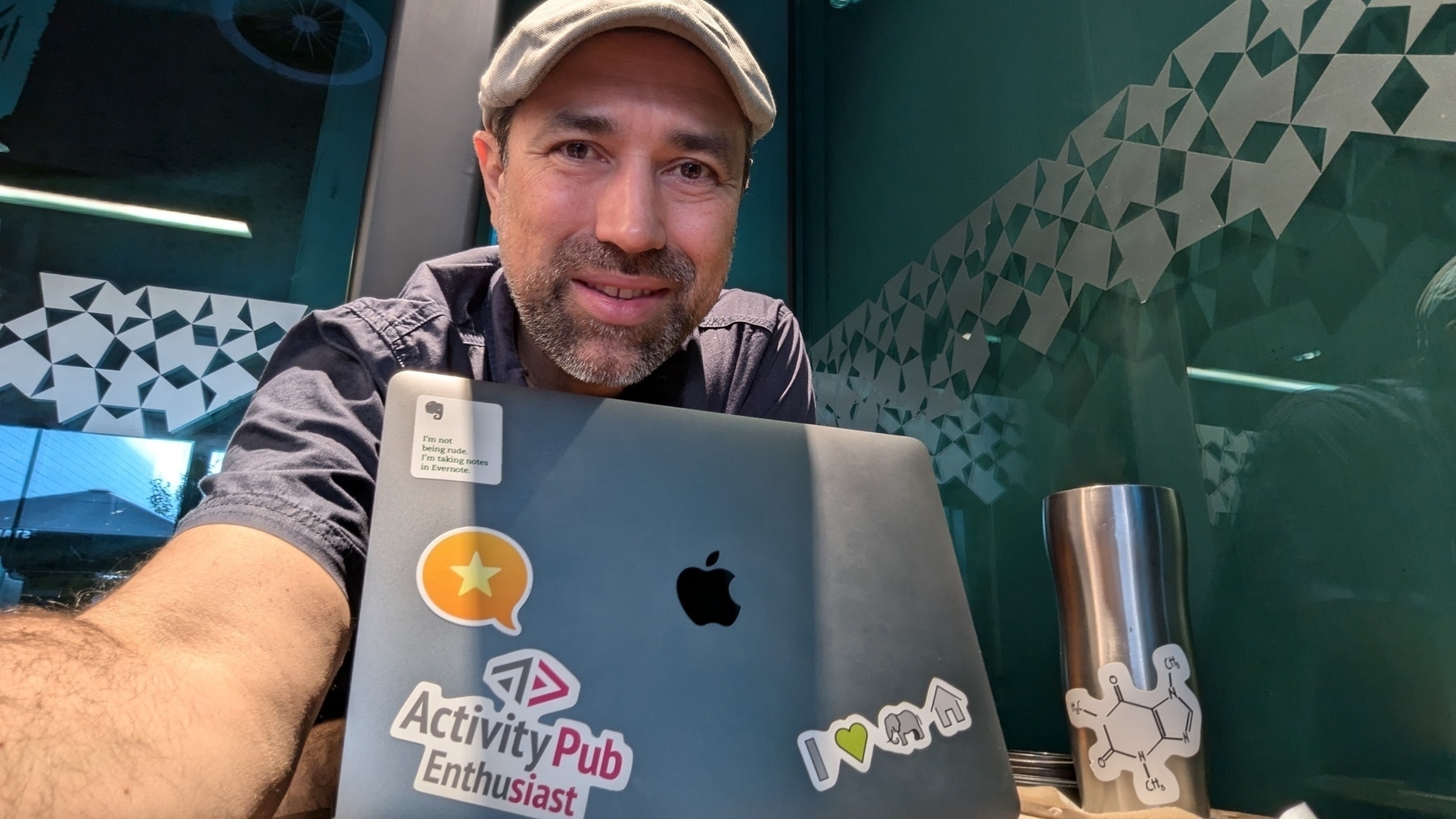 A person with a beard and cap is sitting at a table with a laptop and a drink container covered in various stickers.