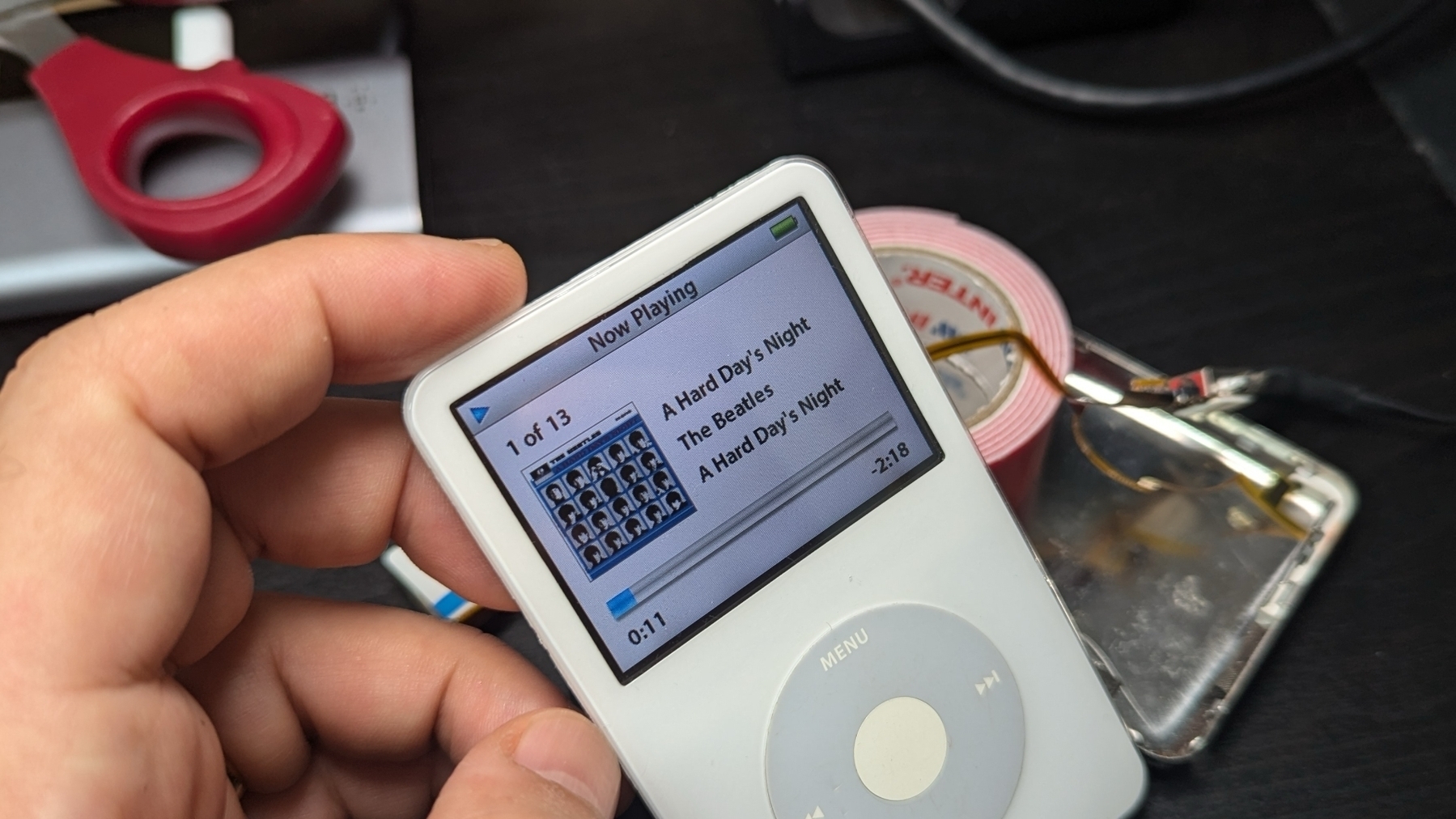 A person is holding a white iPod displaying The Beatles' A Hard Day's Night with various items like a can and wires in the background.